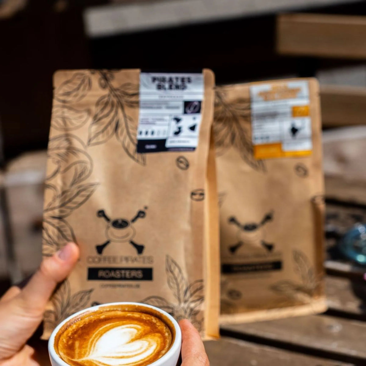 Coffee Pirates Kaffeebeutel im Hintergrund Cappuccino in der Hand